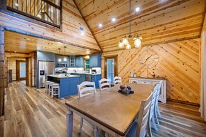 Cozy cabin-style interior with a high ceiling and exposed beams, featuring a central island and dining area with a large wooden table.