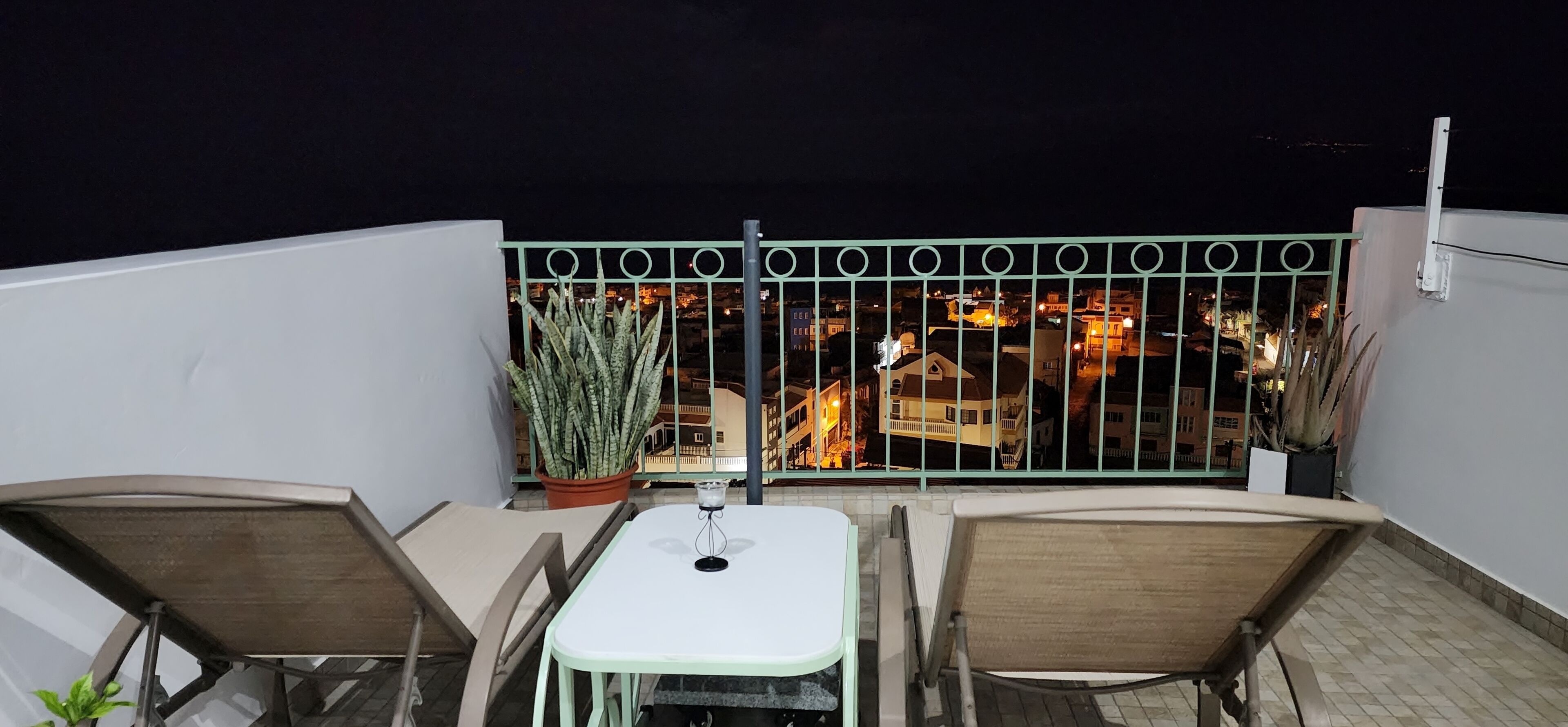 A balcony with two chairs and a table, a potted plant, and a view of a cityscape at night.