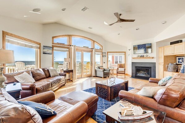 A spacious living room with leather sofas, a fireplace, and large windows offering a view of the city. - Wink Beach | Oceanfront | Private Pool, Hot Tub, and Dog Friendly (Nags Head)