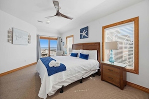 A spacious bedroom with a large bed, a ceiling fan, a wooden nightstand, and a view of the ocean through the window. - Wink Beach | Oceanfront | Private Pool, Hot Tub, and Dog Friendly (Nags Head)