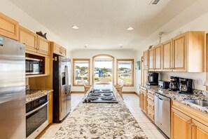 A modern kitchen with wooden cabinets, stainless steel appliances, a granite countertop, and large windows offering a view of the outdoors. - Wink Beach | Oceanfront | Private Pool, Hot Tub, and Dog Friendly (Nags Head)