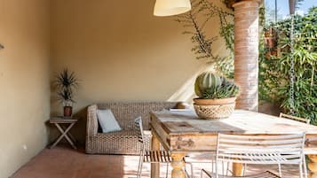 Een terras met een houten tafel en stoelen, een geweven bank, een bijzettafeltje met een plant en een hanglamp.