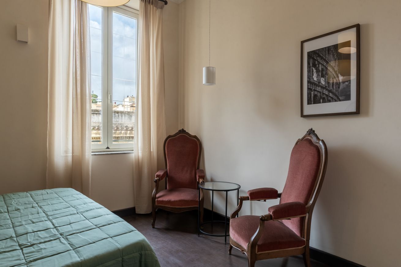 Une chambre d’hôtel avec un lit, deux fauteuils rouges, une petite table ronde, une fenêtre avec des rideaux et un tableau encadré au mur.
