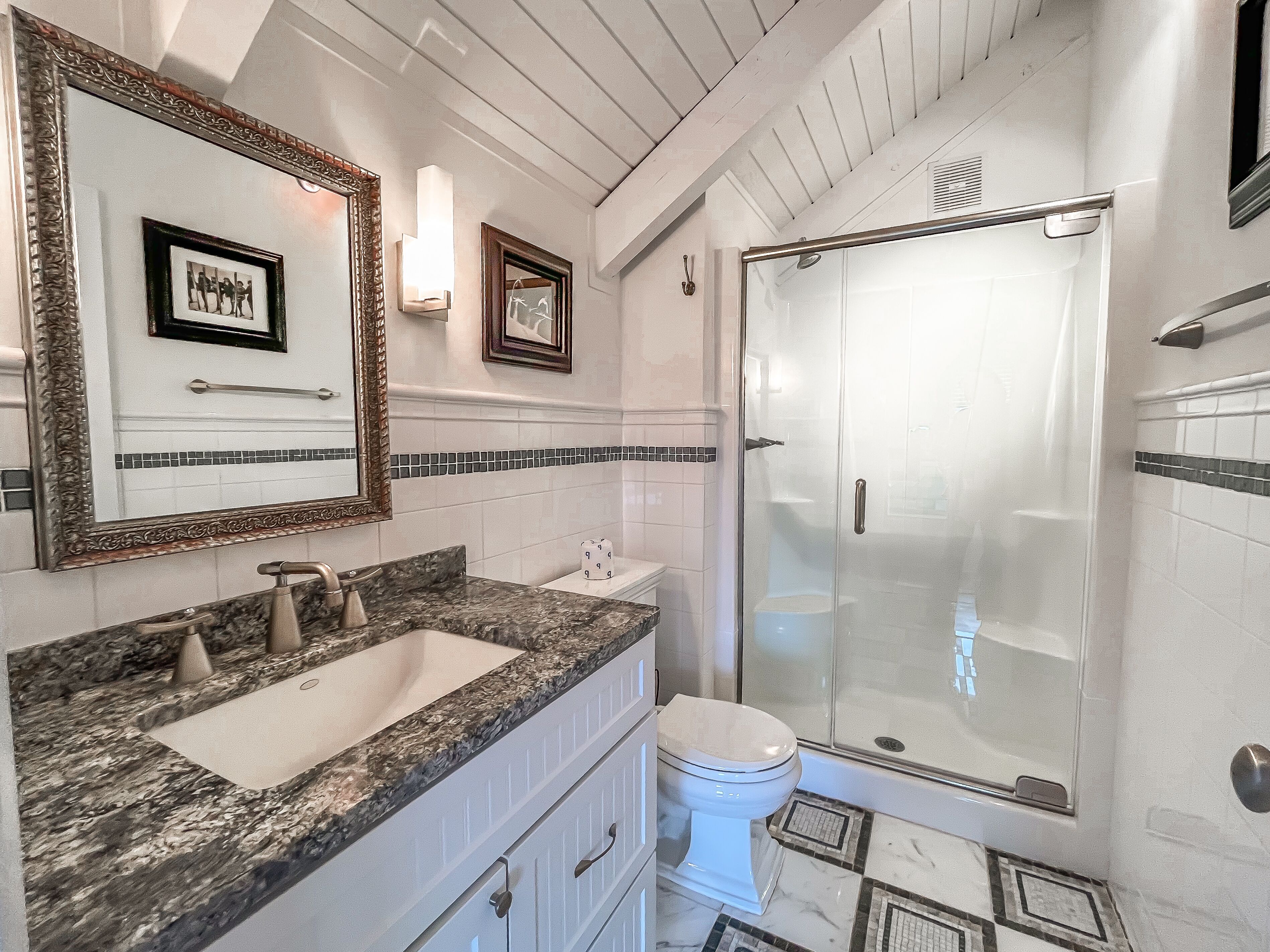 A bathroom with a granite countertop sink, a toilet, a glass-enclosed shower, and two framed pictures on the wall.
