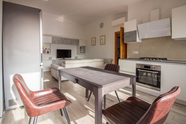 A modern kitchen with a dining table and chairs, a built-in oven, and a television in the living area.