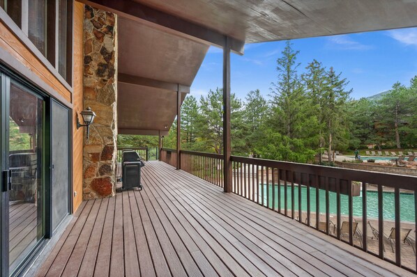 A wooden deck with a stone wall, a grill, and a view of a swimming pool and trees. - Red Pine Y4 | Mountain Views | Fireplace | Kitchen | Patio | Grill | Pools & Hot Tub | Walk to Lifts (Park City)