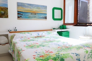 A bedroom with a bed, a painting of a beach scene, a green mirror, and a window with blinds. - GuestHost - Comfy Flat W/ Pool, Tennis Court & Private Beach! (Pischina Salida)