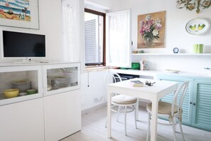 Eine Küche mit weißen Schränken, einem Esstisch, Stühlen, einem Fernseher und einem Fenster mit Jalousien.