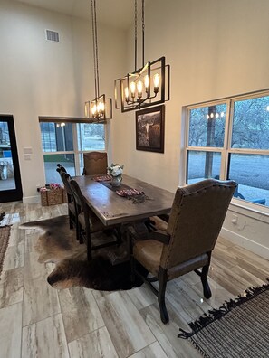 A dining area with a long wooden table, multiple chairs, hanging pendant lights, and a large window with a view of a snowy landscape. - Perfect Couple’s Getaway in Luckenbach Tx Hot tub Pool (Fredericksburg)