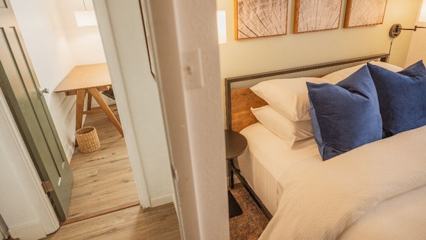 A bedroom with a bed, pillows, a bedside table, and a lamp. There is a door leading to another room and a small table with a lamp in the corner.