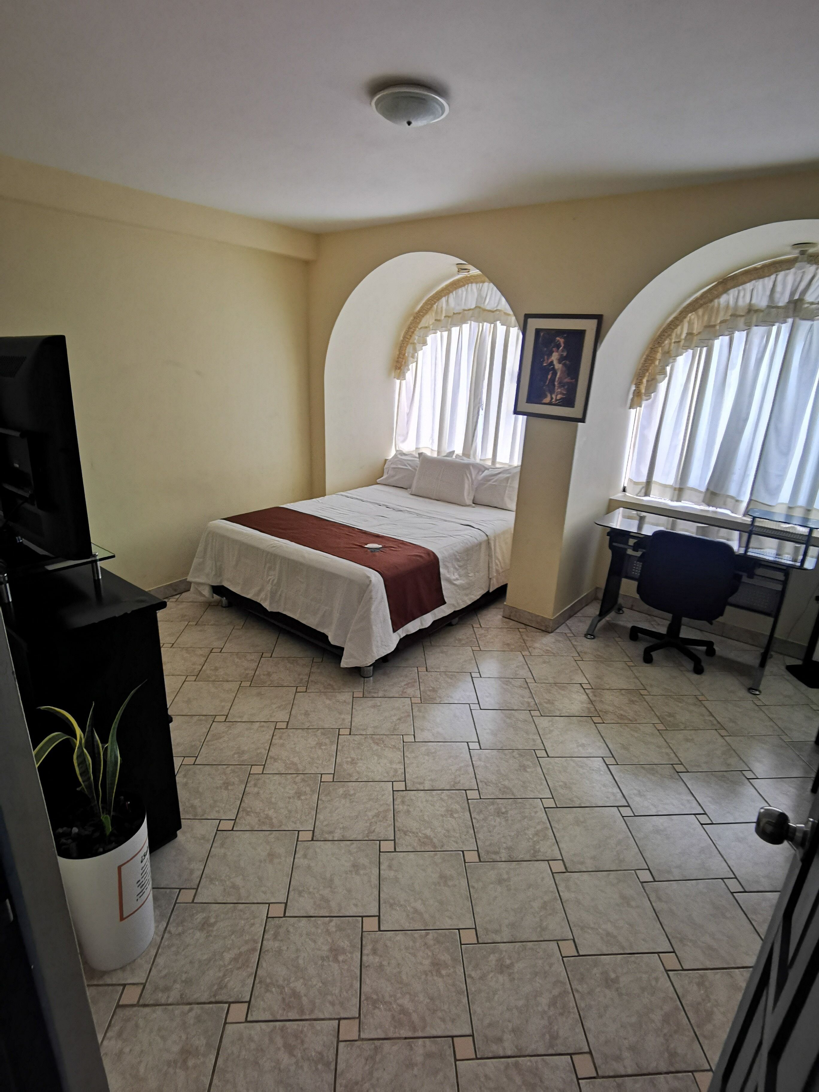 A bedroom with a bed, a desk, and a chair. There is a window with curtains and a ceiling light.