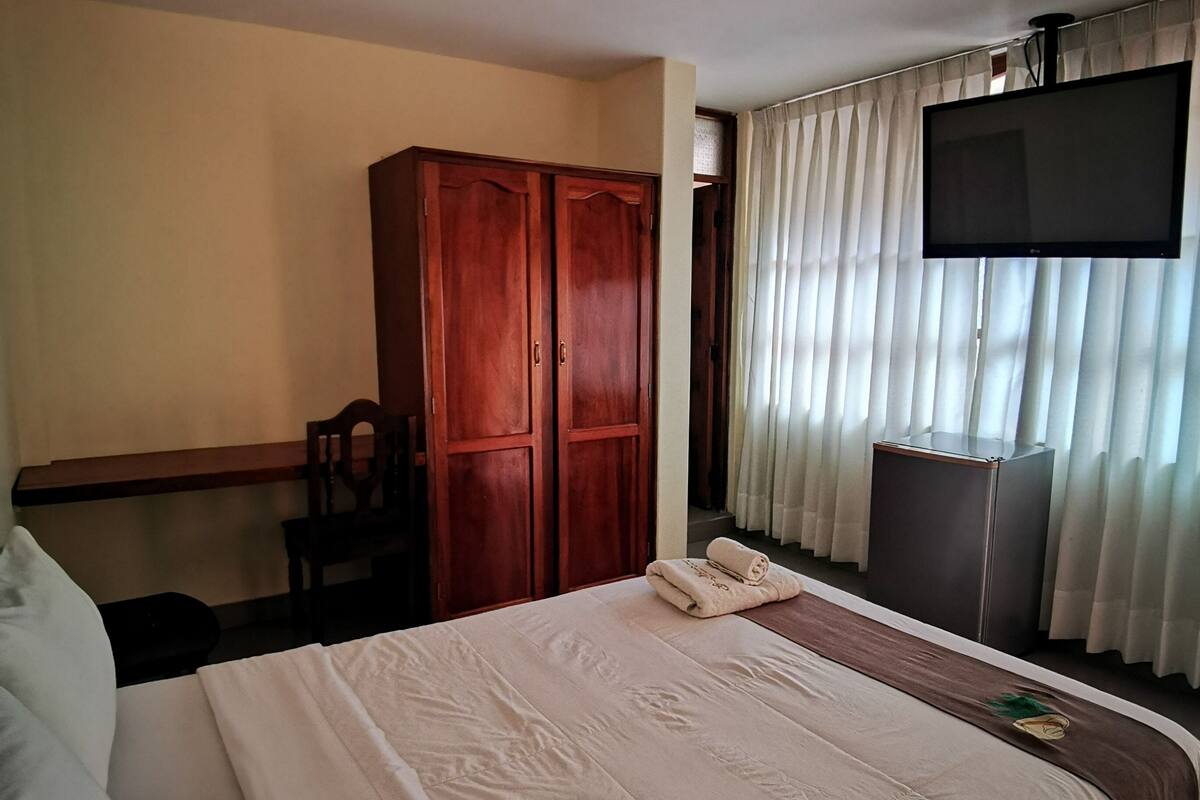 A hotel room with a large bed, a wardrobe, a desk, a chair, a television, and a window with curtains.