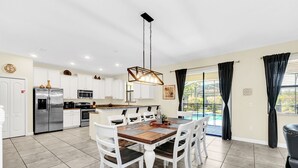 A modern kitchen with white cabinets, a stainless steel refrigerator, and a dining area with a wooden table and white chairs. - Splendor Retreat| 6BR Villa w/ Kid's Room & Pool (Davenport)