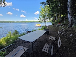 Un área al aire libre junto al lago con mesa y sillas, una parrilla y un muelle flotante con un bote inflable.