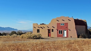 A single-story building with a red door and windows, surrounded by dry grass and shrubs, with mountains in the background.