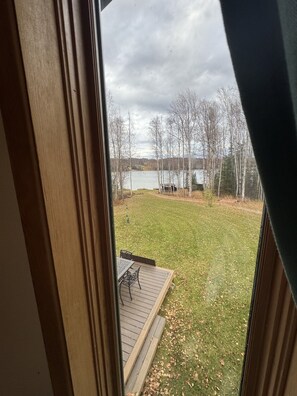 Vista desde una ventana que muestra un patio trasero con una terraza de madera, un césped y un lago al fondo.