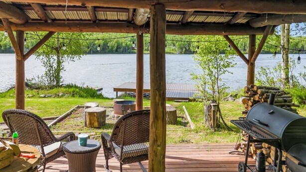 Un espace extérieur abrité, équipé d’un barbecue, de chaises et d’une table, avec vue sur un lac.