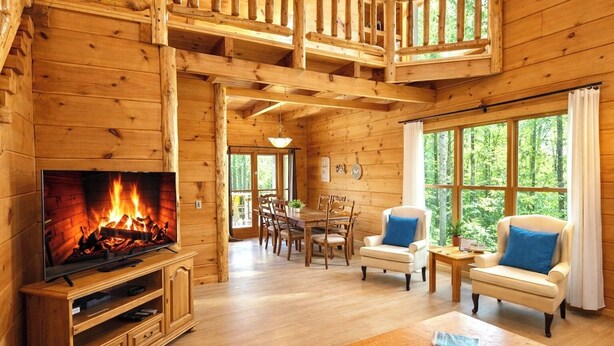 A wooden cabin interior with a TV displaying a fireplace, a dining area, and a seating area with two armchairs and a table. - Eagle Lake Cabin Coalmont (Coalmont)