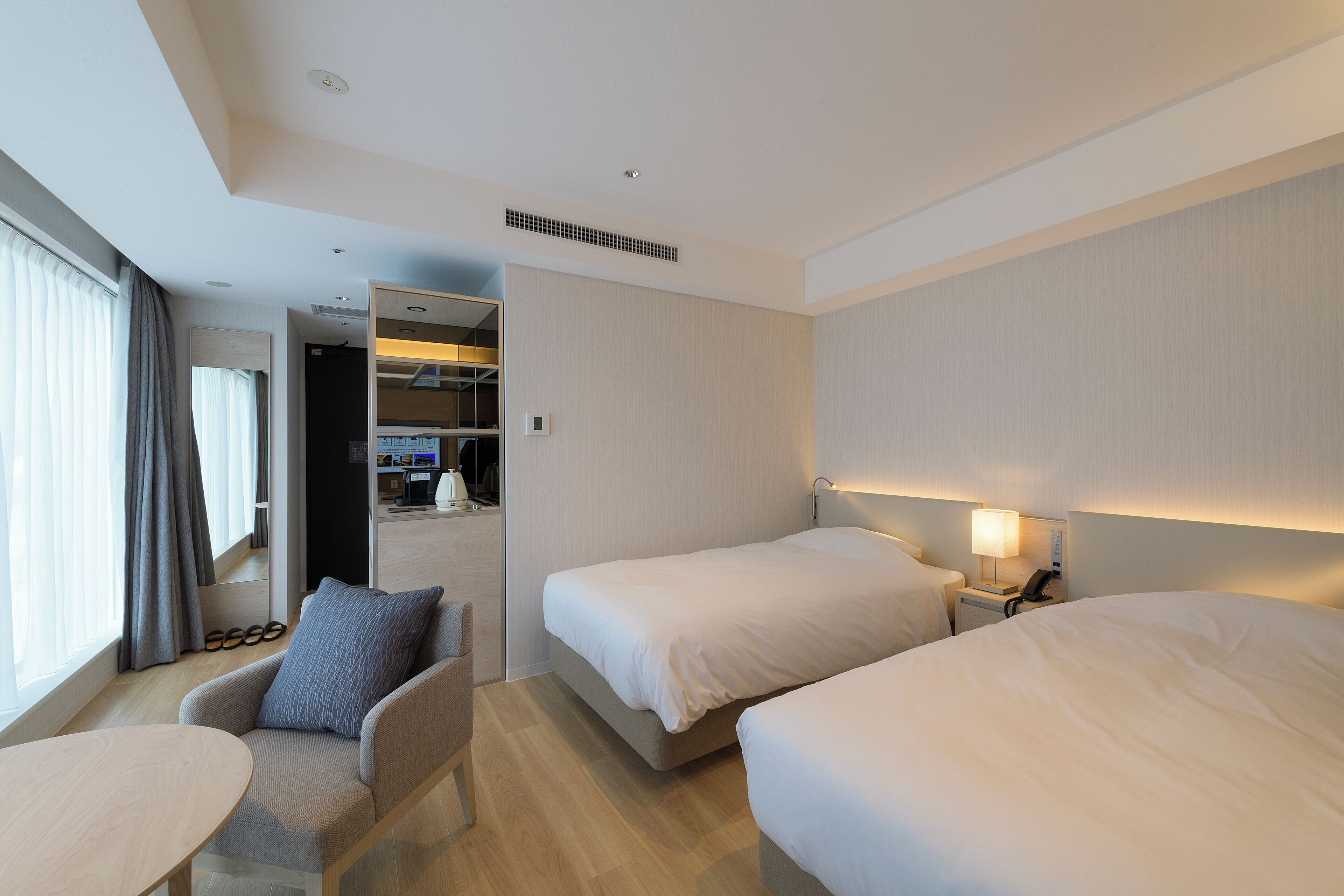 Habitación de hotel con dos camas, un escritorio, una silla y un amplio ventanal con cortinas.
