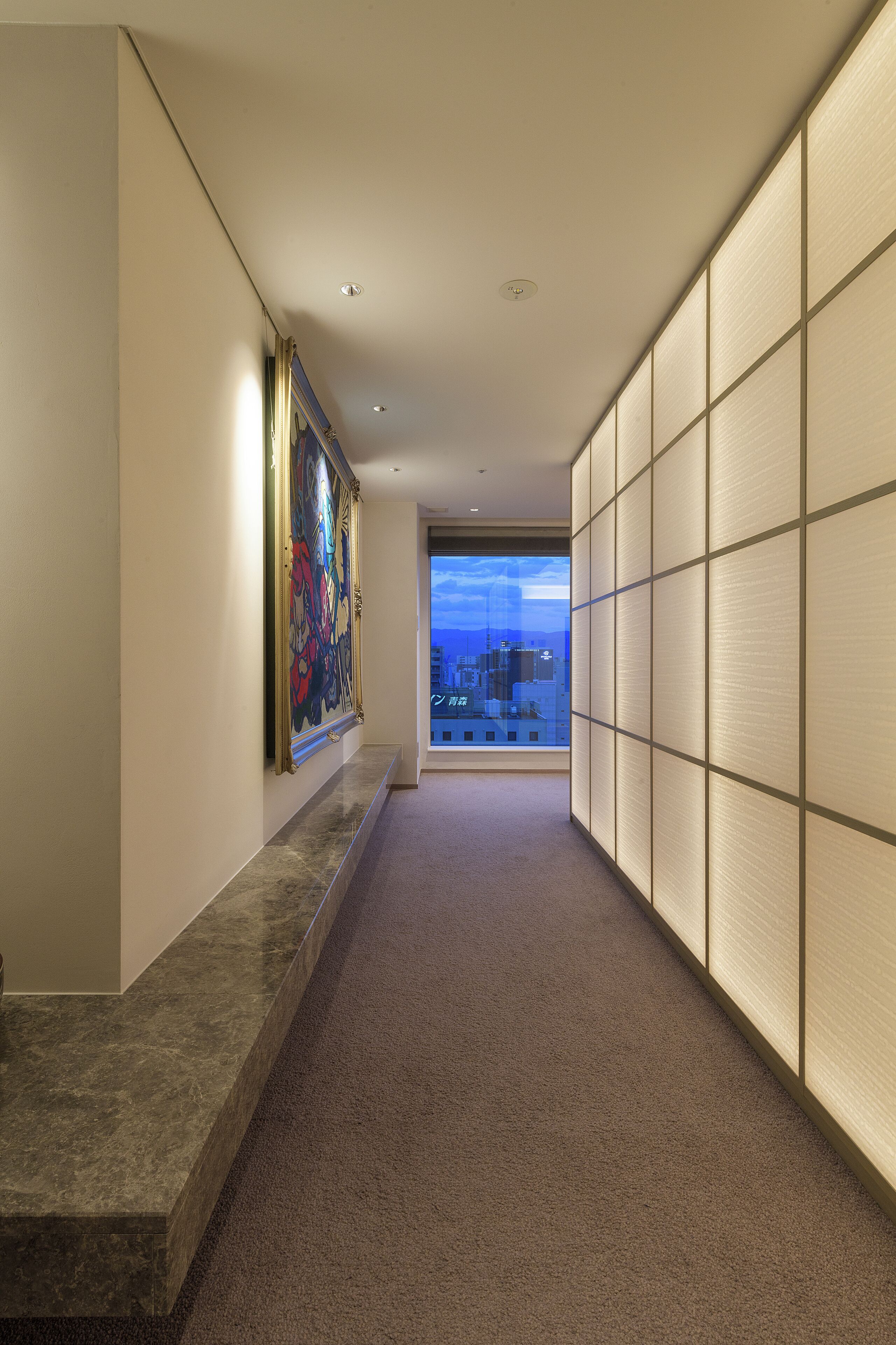A hallway with a marble floor, a large framed artwork on the left, and a view of the city through a large window.