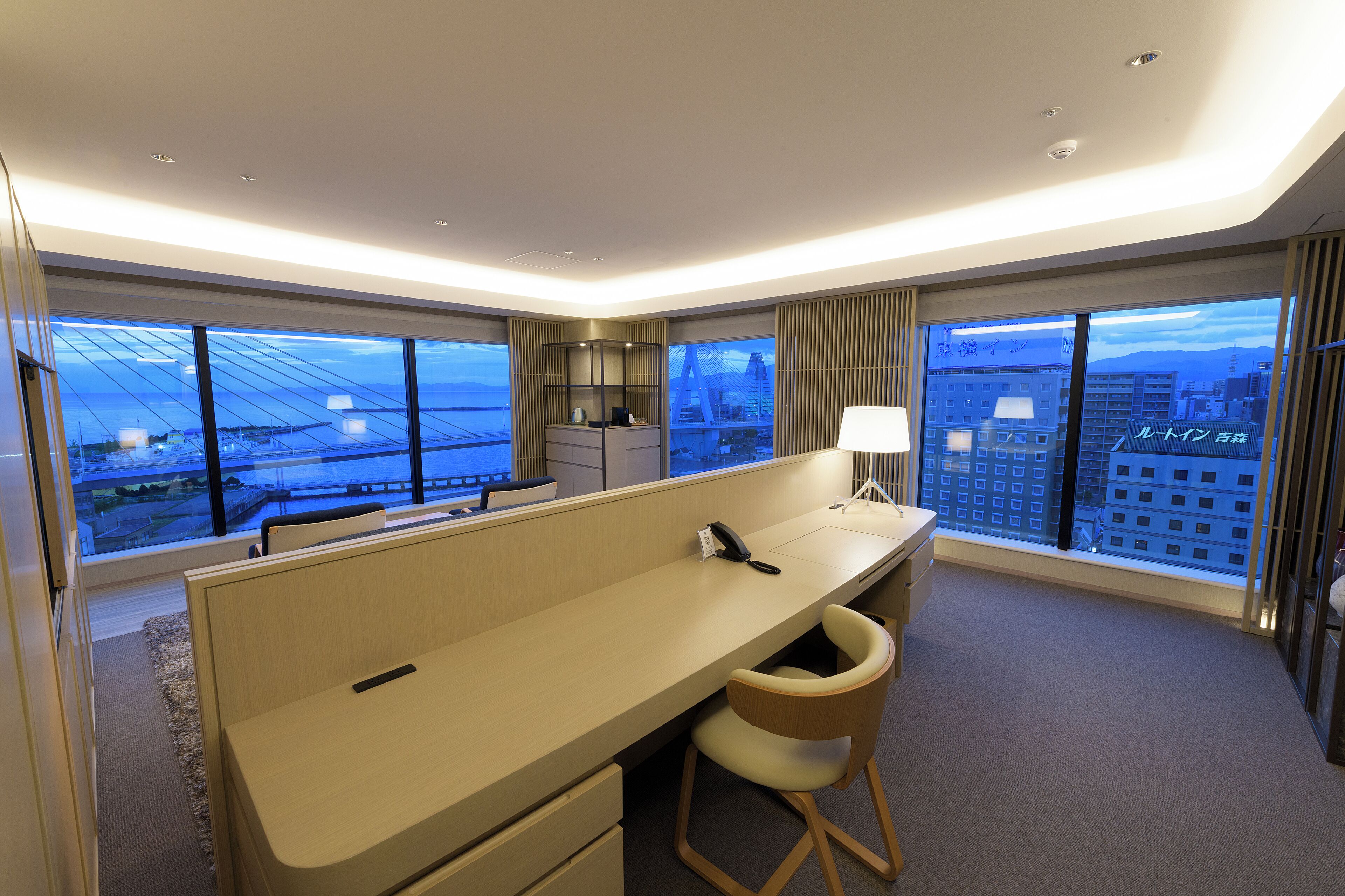 A modern hotel room with a large desk, a chair, a phone, and a lamp. There are large windows with a city view.