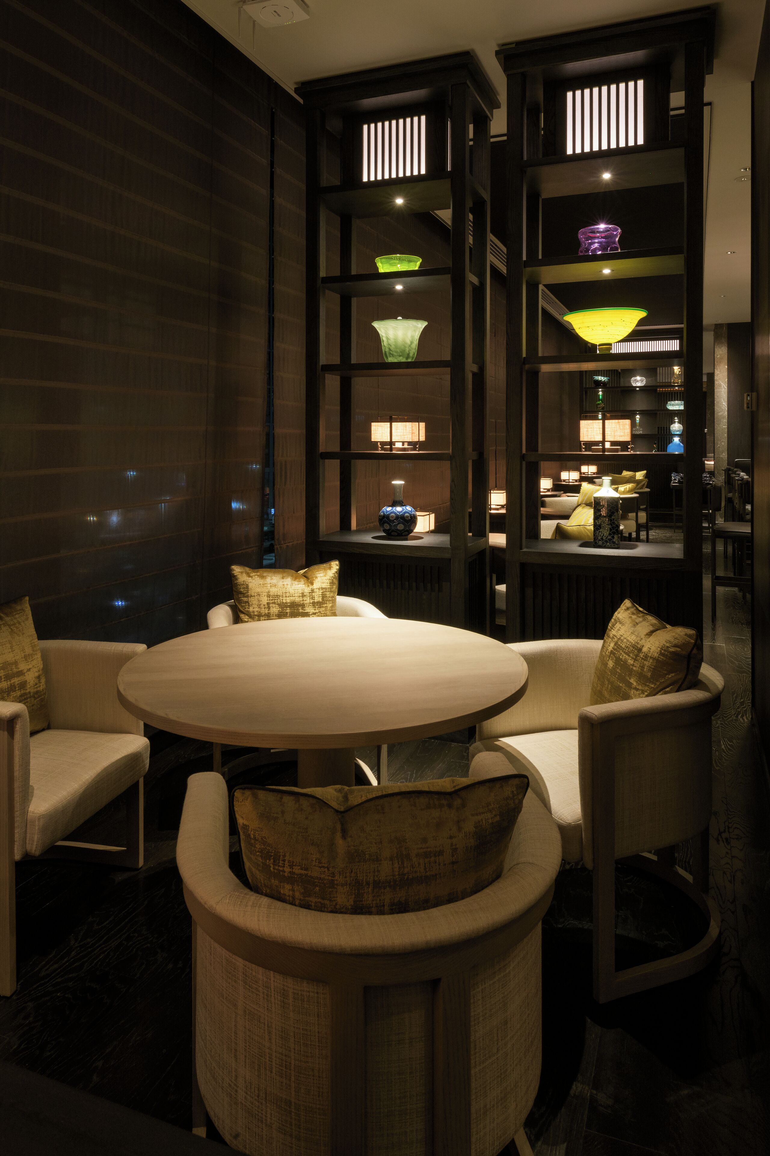 A modern dining area with a round table and cushioned chairs, surrounded by dark wooden paneling and illuminated by various hanging lamps.