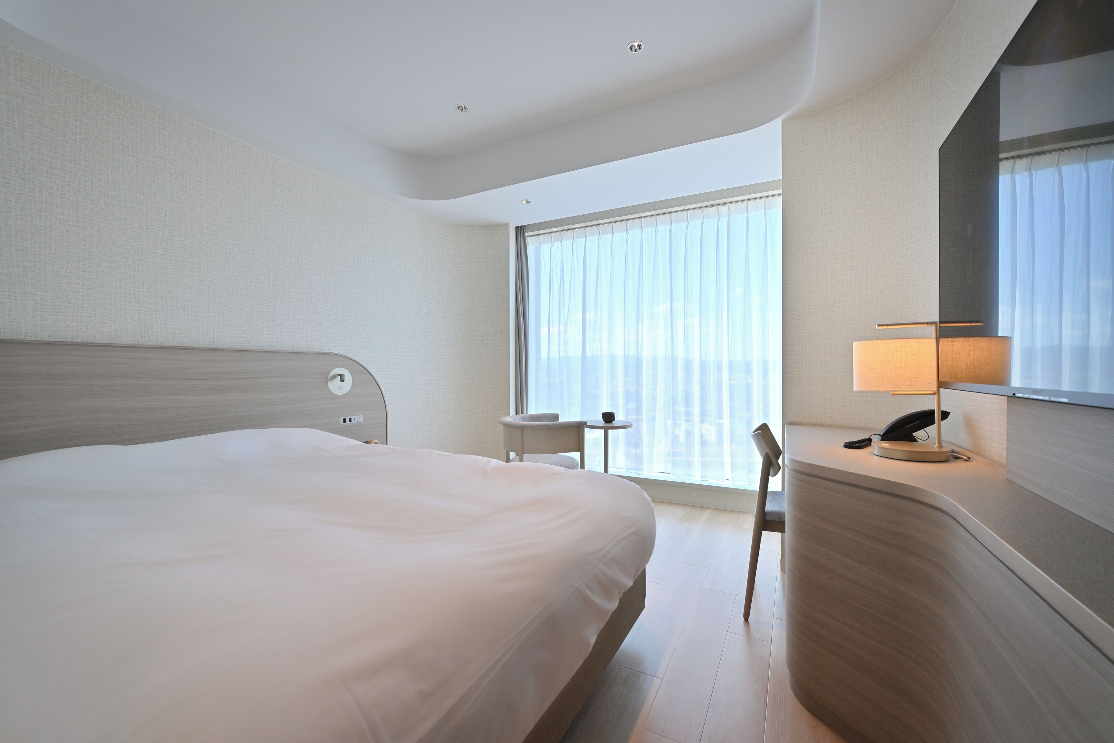 A hotel room with a large bed, a desk with a lamp, a chair, and a view of the city through large windows.