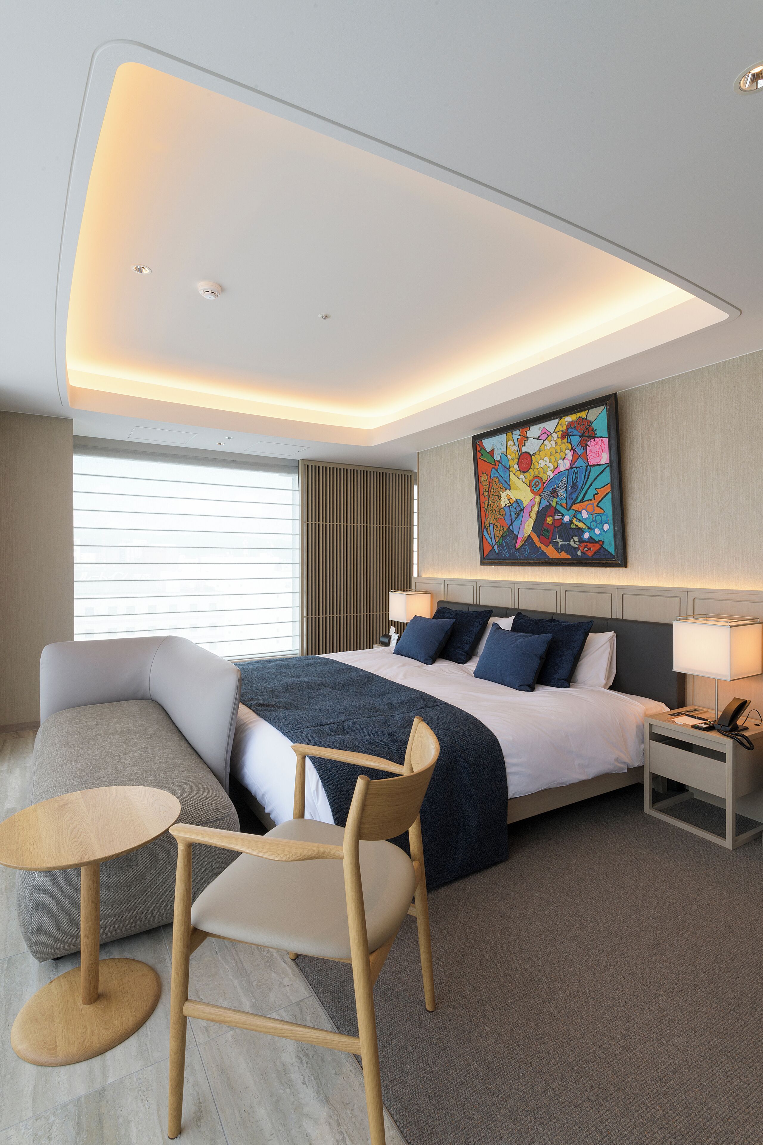 Une chambre d’hôtel moderne dotée d’un grand lit, d’un coin salon avec un fauteuil et une table, et d’une grande fenêtre avec des stores.