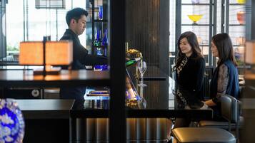 A bar area with a bartender, a customer, and a server. There are shelves with decorative items and a large window with natural light.