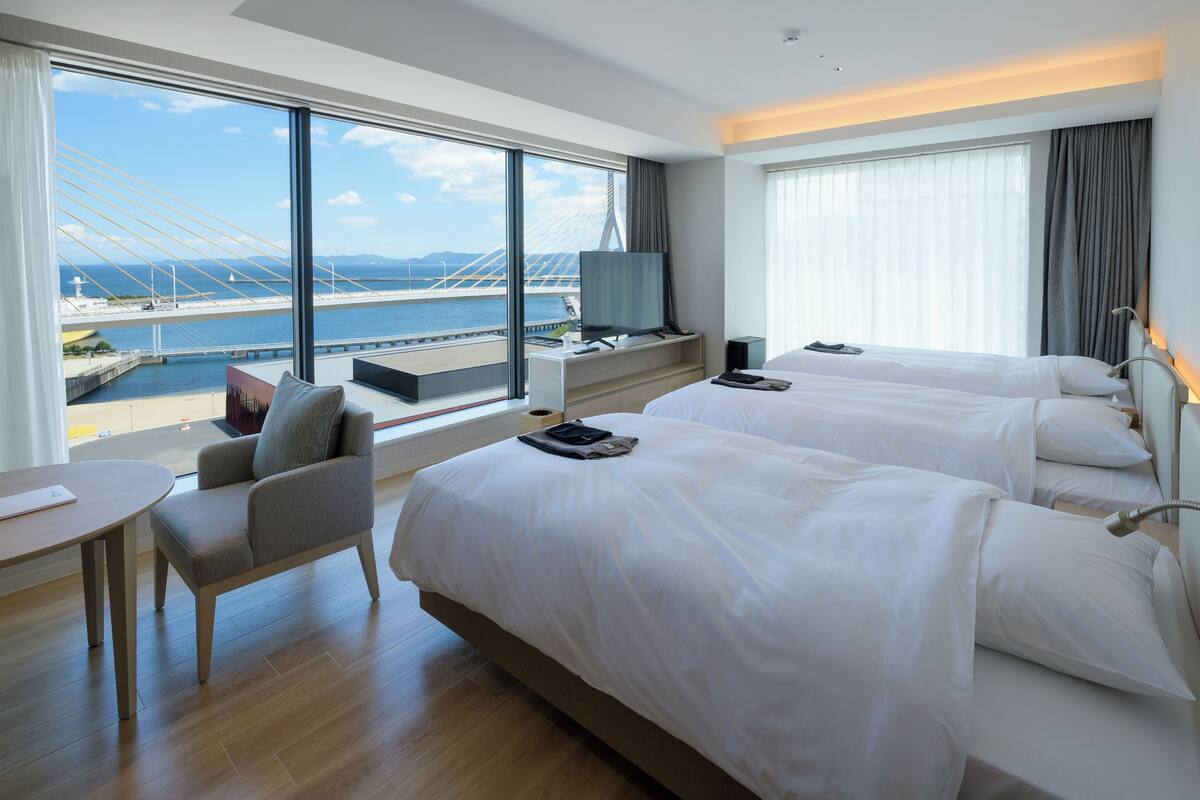 Une chambre d’hôtel avec deux lits, un bureau, une chaise et une grande fenêtre donnant sur un pont et sur l’eau.