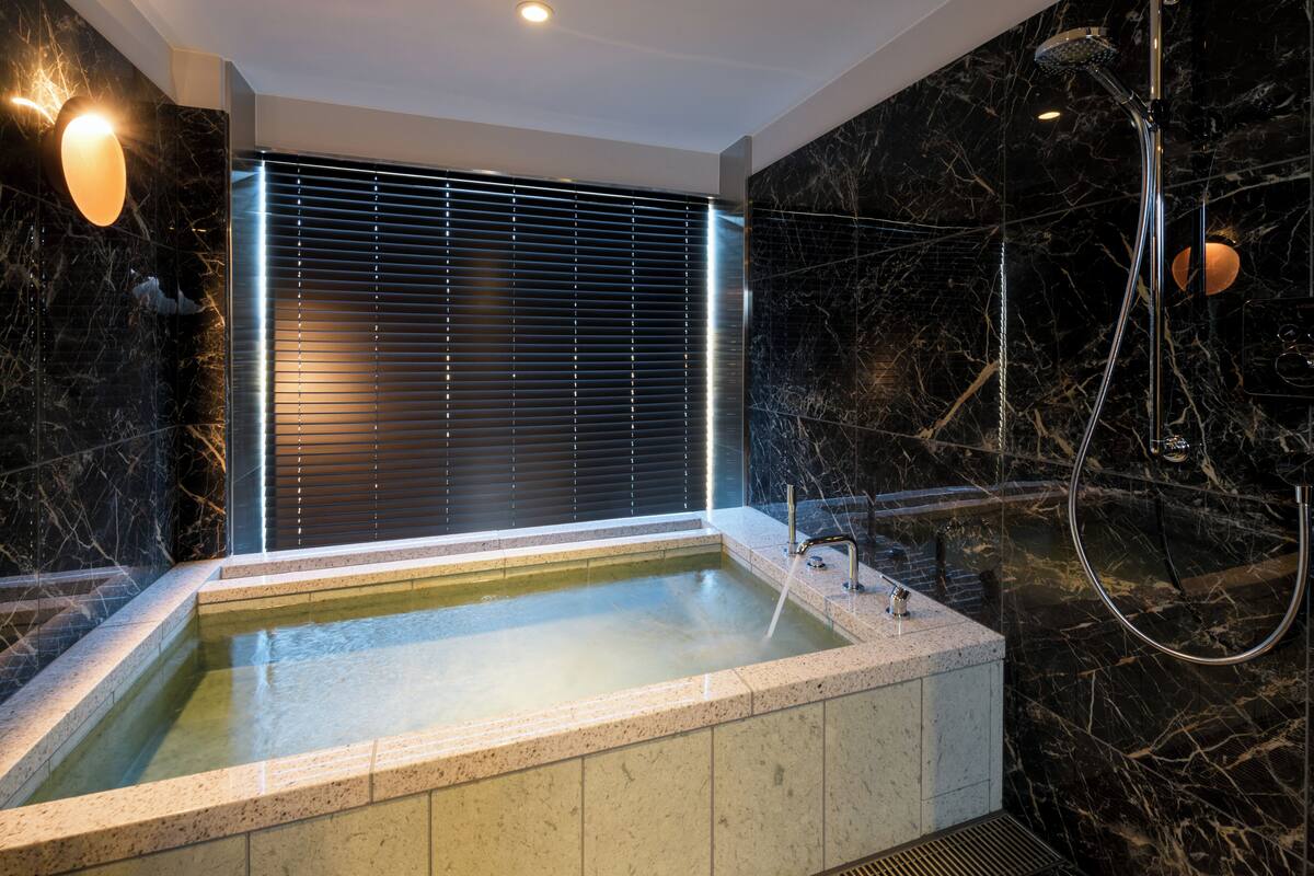 Une salle de bain moderne avec un mur en marbre, une baignoire intégrée, un espace douche et une fenêtre avec des stores.