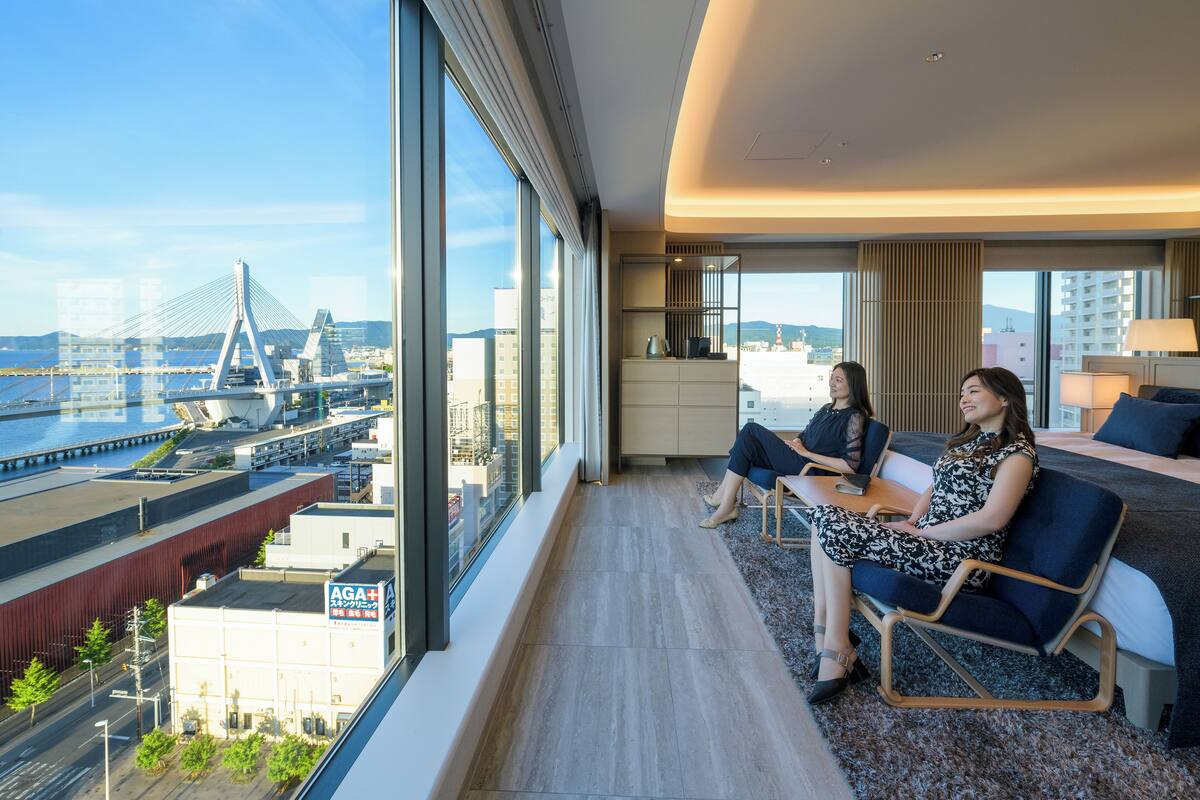 Une chambre d’hôtel moderne dotée de grandes fenêtres, offrant une vue sur la ville, un balcon et une vue sur un pont.