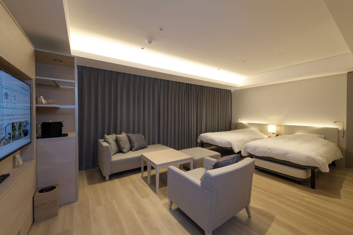 Une chambre d’hôtel moderne avec un canapé, deux lits simples, une petite table et un téléviseur posé sur un meuble.