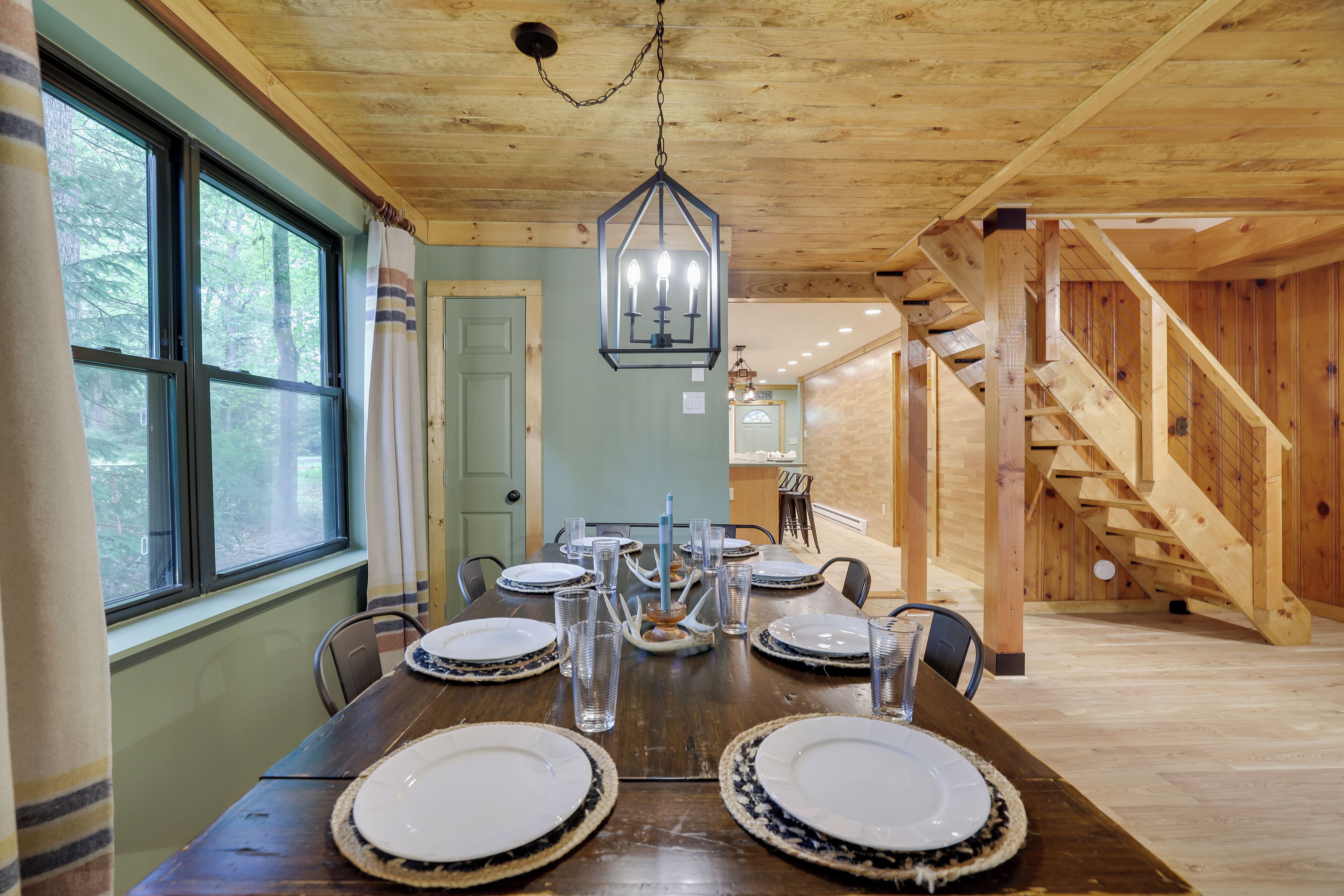 Un espace repas avec une table en bois dressée pour six personnes, un lustre et un escalier menant à un niveau supérieur.