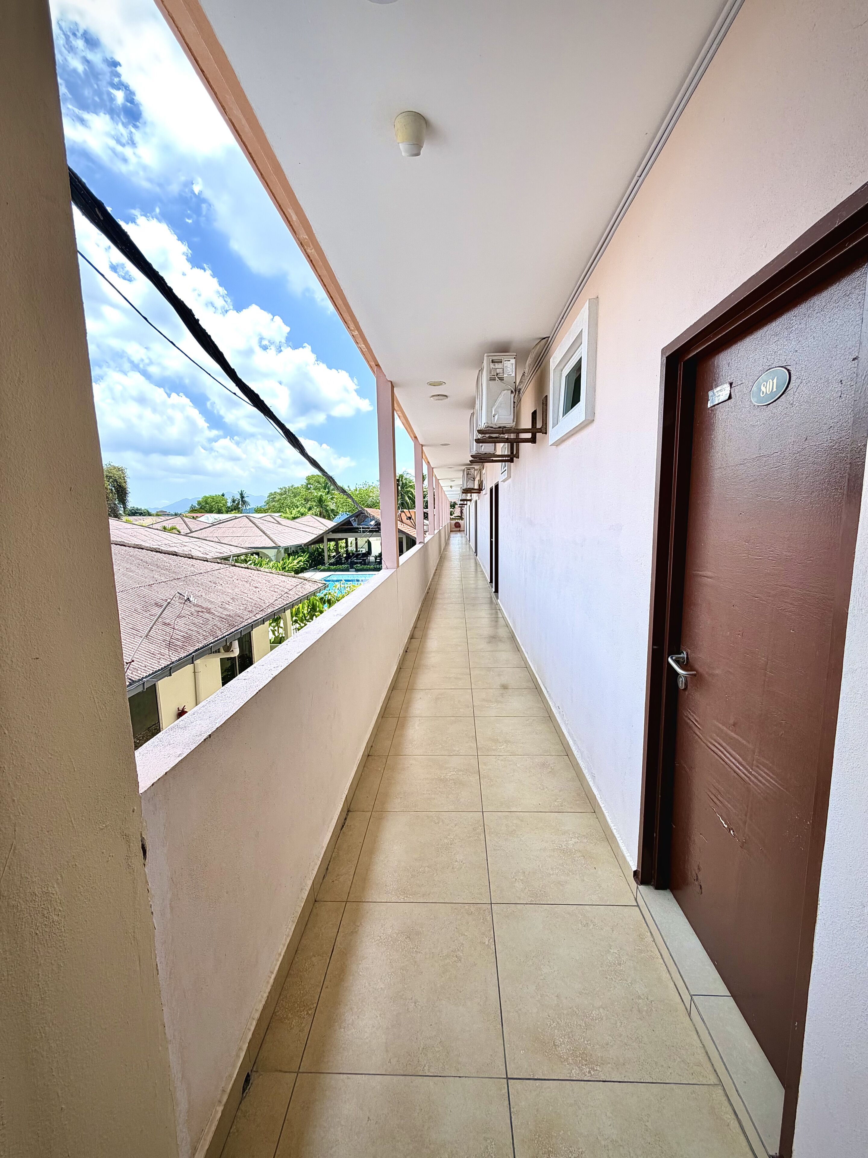 A corridor with a tiled floor, a brown door with a number plate, and air conditioning units on the wall.