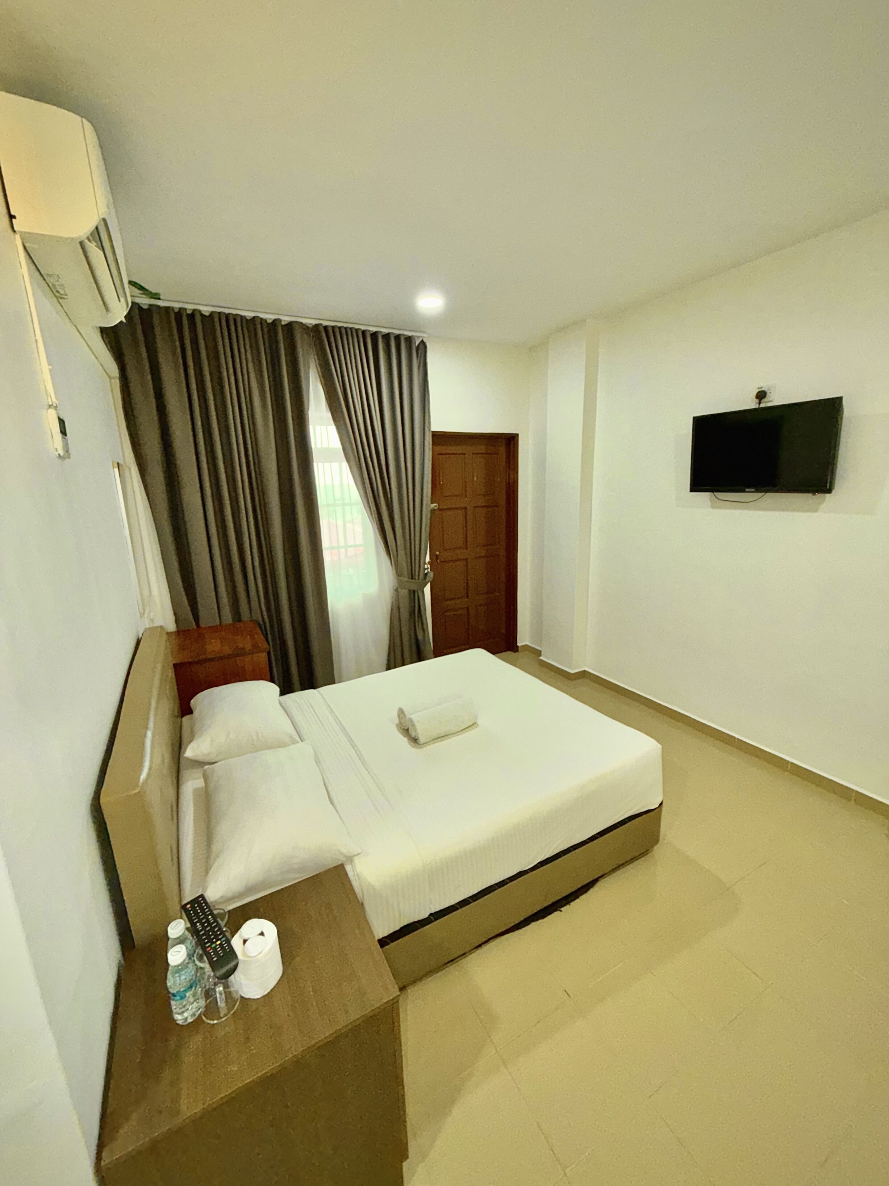 Habitación de hotel con una cama, un televisor montado en la pared, un cabecero de madera, una ventana con cortinas y una mesita con control remoto y botellas de agua.