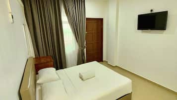 Habitación de hotel con una cama, un televisor montado en la pared, un cabecero de madera, una ventana con cortinas y una mesita con control remoto y botellas de agua.