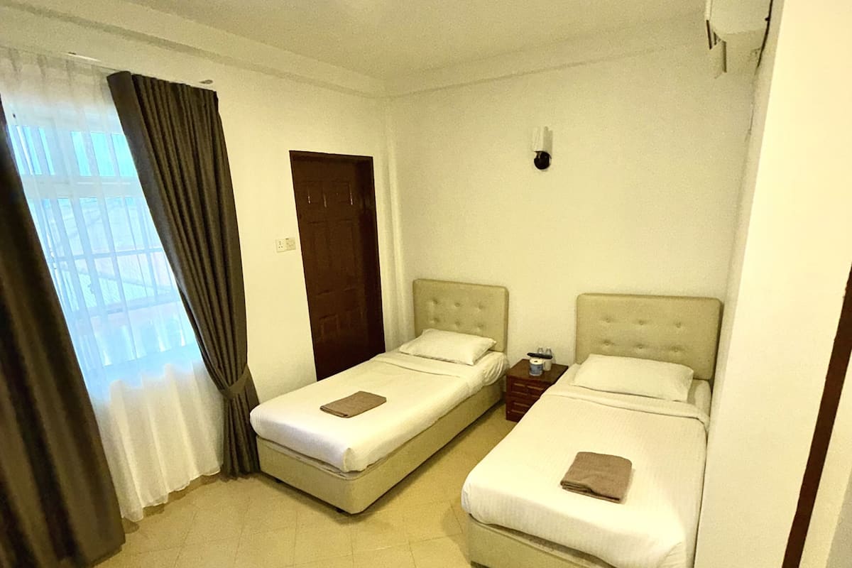 A hotel room with two single beds, a small table, a chair, a window with curtains, and a door.