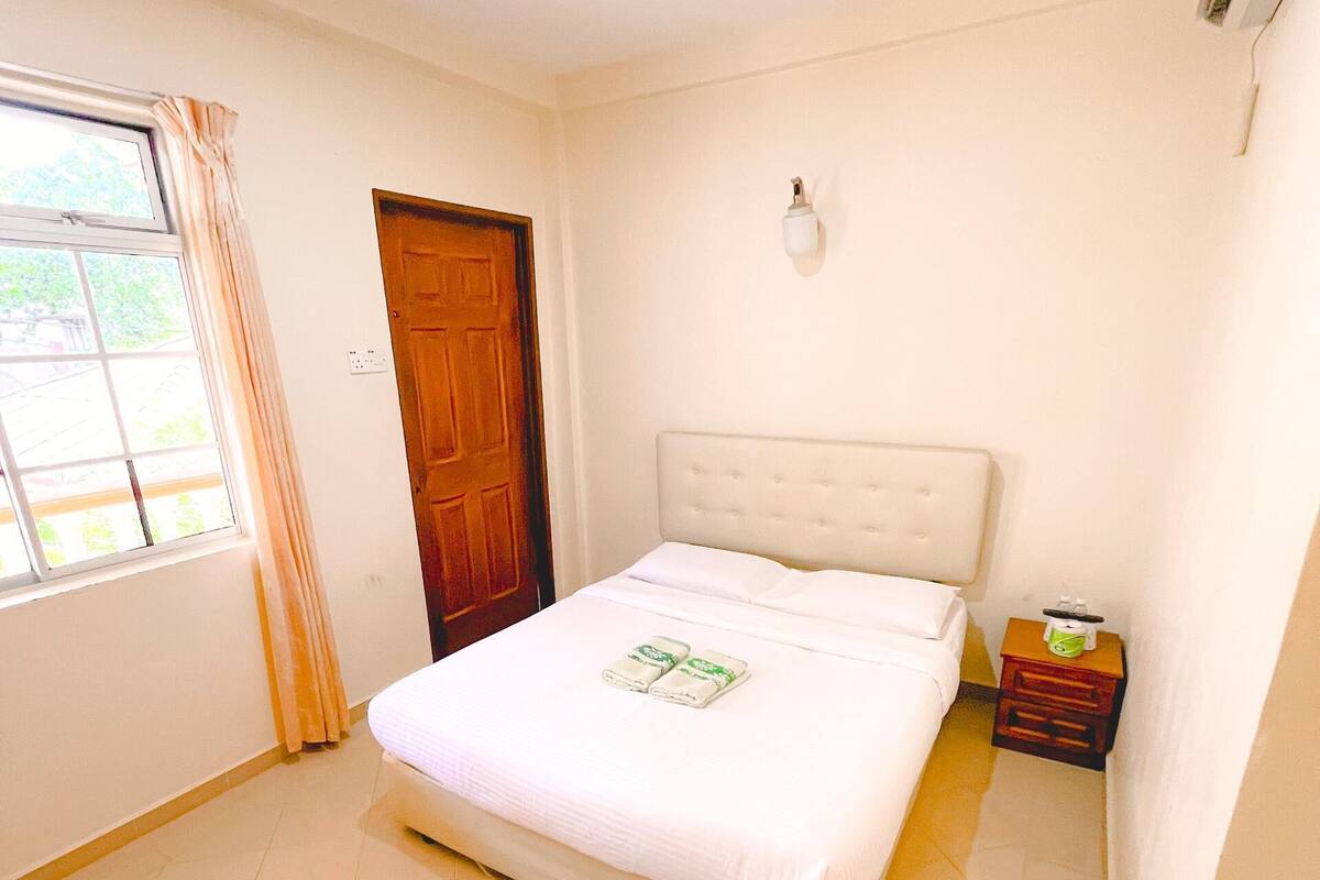 A bedroom with a bed, a wooden door, a window with curtains, and a bedside table with a lamp.