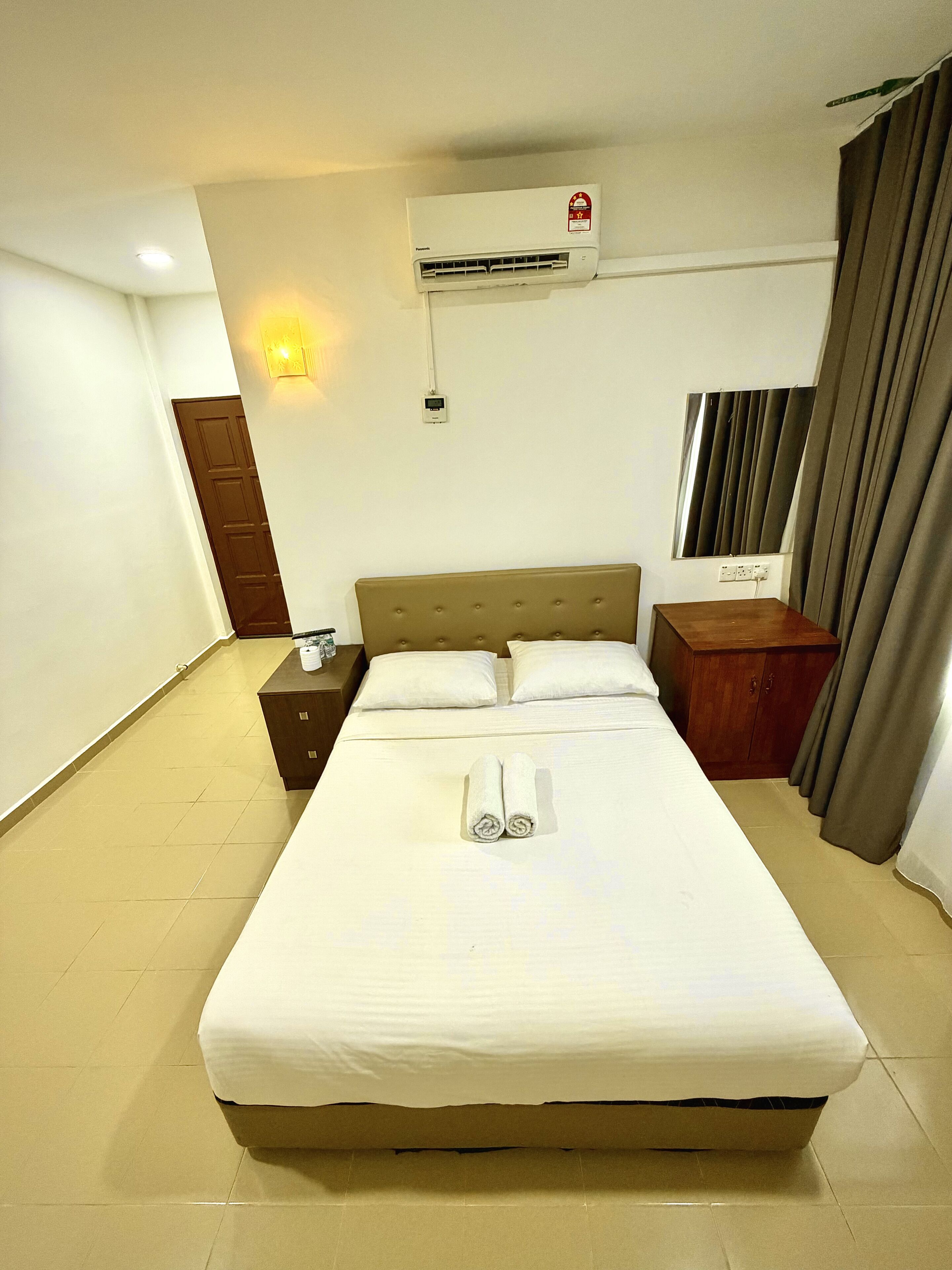 Una habitación de hotel con una cama, una mesita de noche, un aire acondicionado, una lámpara de pared y una ventana con cortinas.