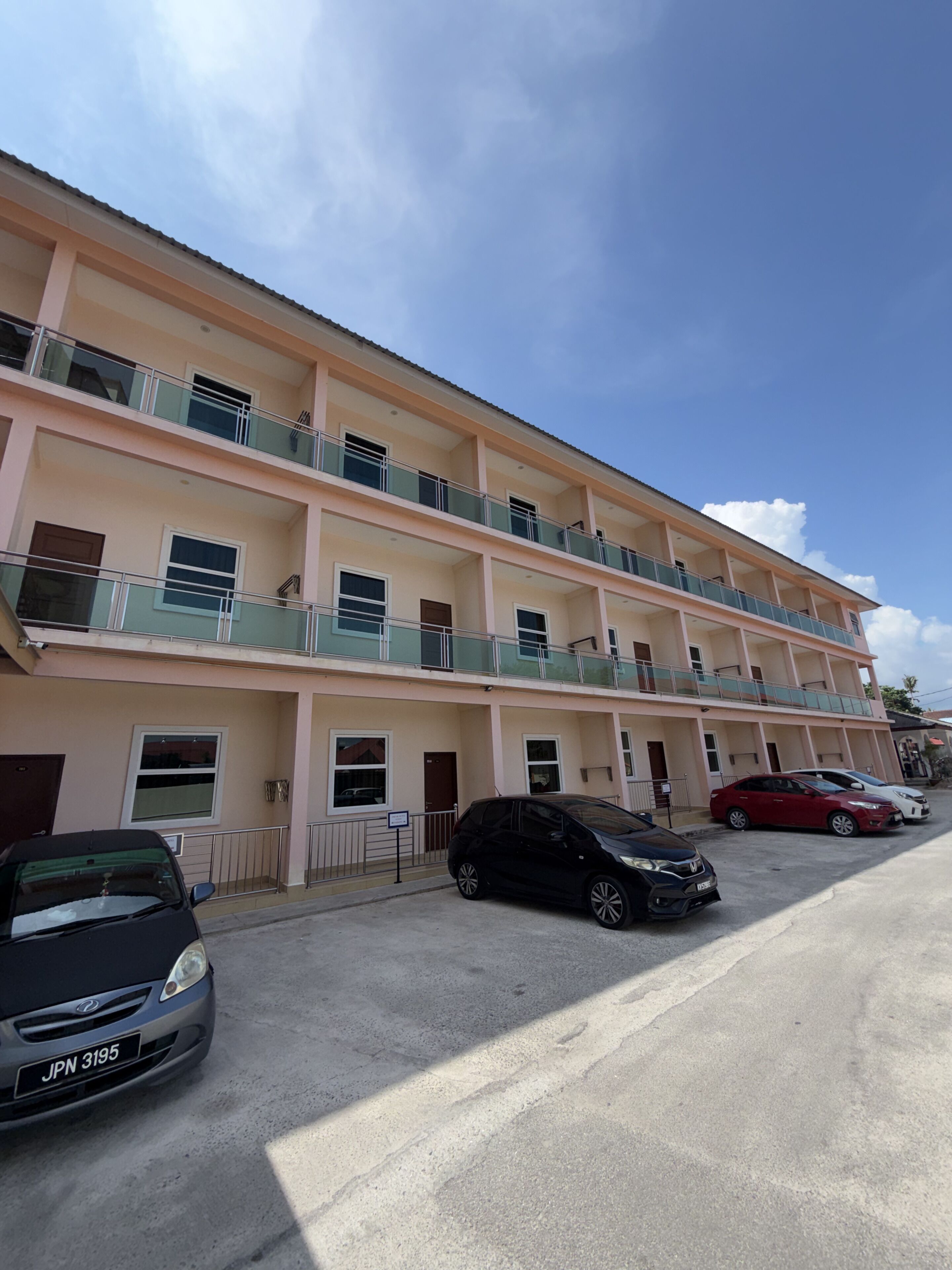 Un edificio de varios pisos con balcones, un estacionamiento con coches y un cielo despejado.