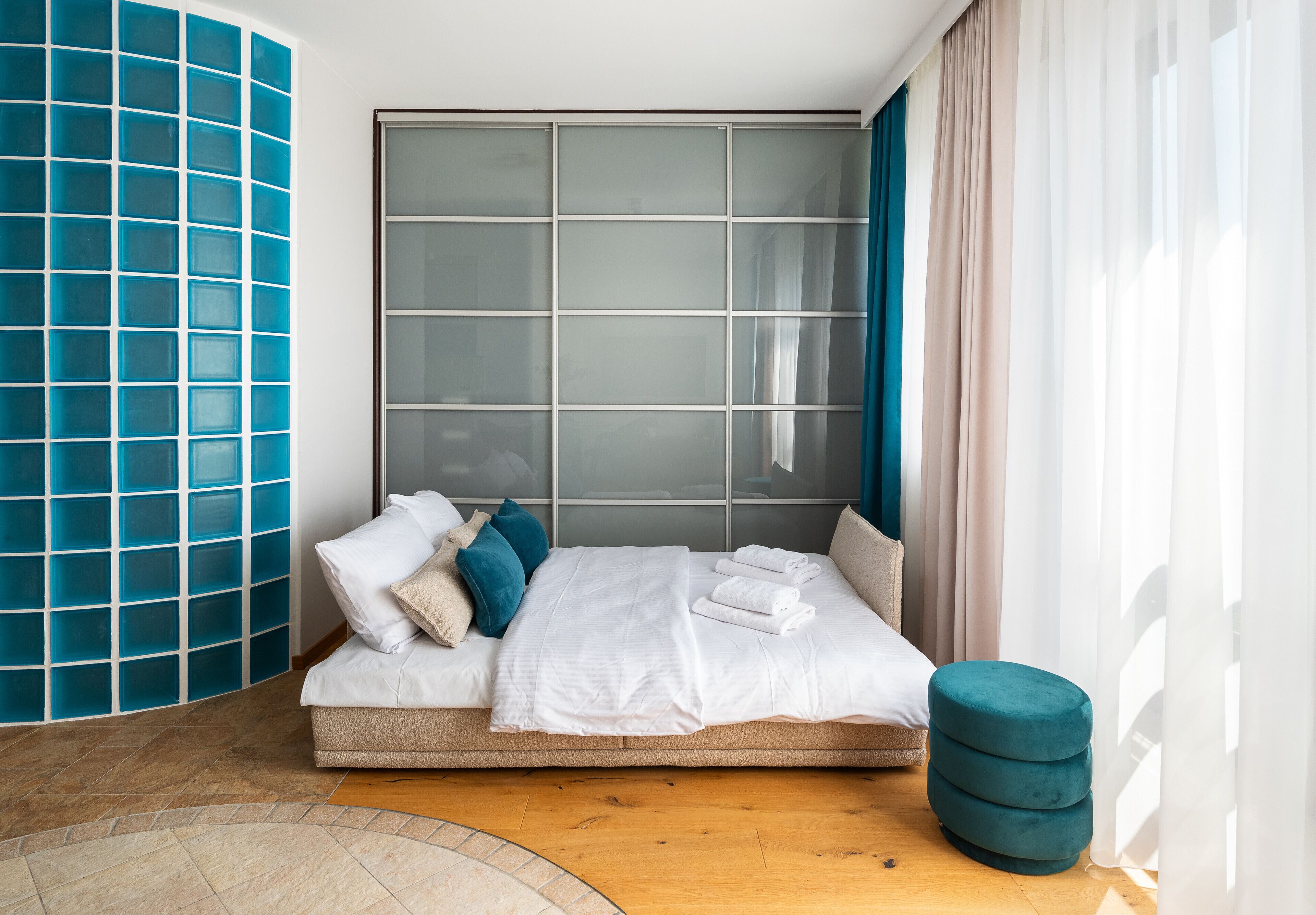 A modern bedroom with a large window, a bed with white linens, a teal ottoman, and a blue tiled accent wall.
