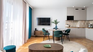 A modern living room with a round wooden coffee table, two green chairs, a dining table with chairs, a flat-screen TV mounted on the wall, and a kitchen area with white cabinets and a wooden countertop. - Saska Kępa Cosy Studio (Warszawa)