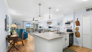 A modern kitchen with a central island, pendant lights, and a dining area with a blue chair.