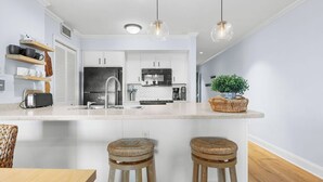 A modern kitchen with white cabinets, a central island with stools, pendant lighting, and a potted plant.