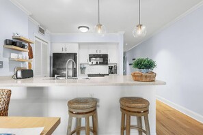 A modern kitchen with white cabinets, a central island with stools, pendant lighting, and a potted plant.