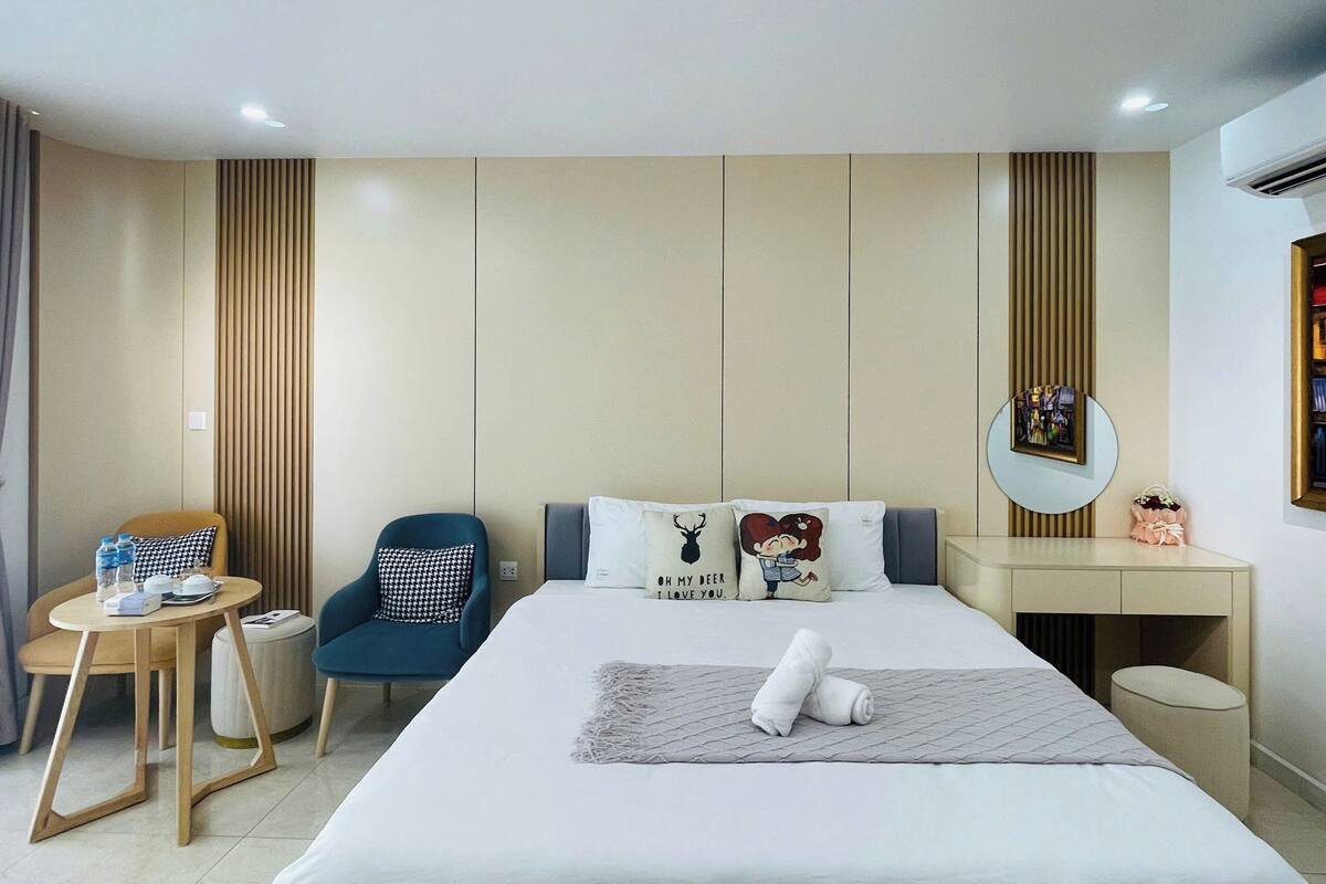 A modern hotel room with a bed, a desk, a chair, a small table with a water bottle, and a mirror on the wall.