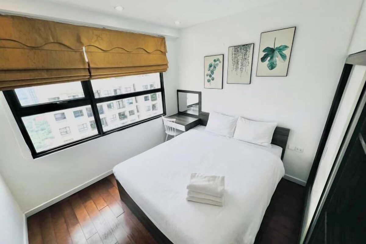 A hotel room with a large bed, a bedside table, a window with a view of buildings, and three framed pictures on the wall.