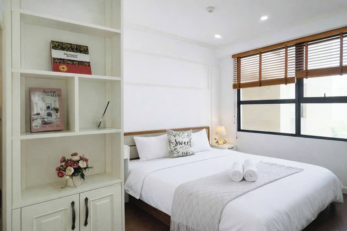 A neatly arranged bedroom with a bed, a bedside table with a lamp, a window with blinds, and a white wardrobe with shelves.