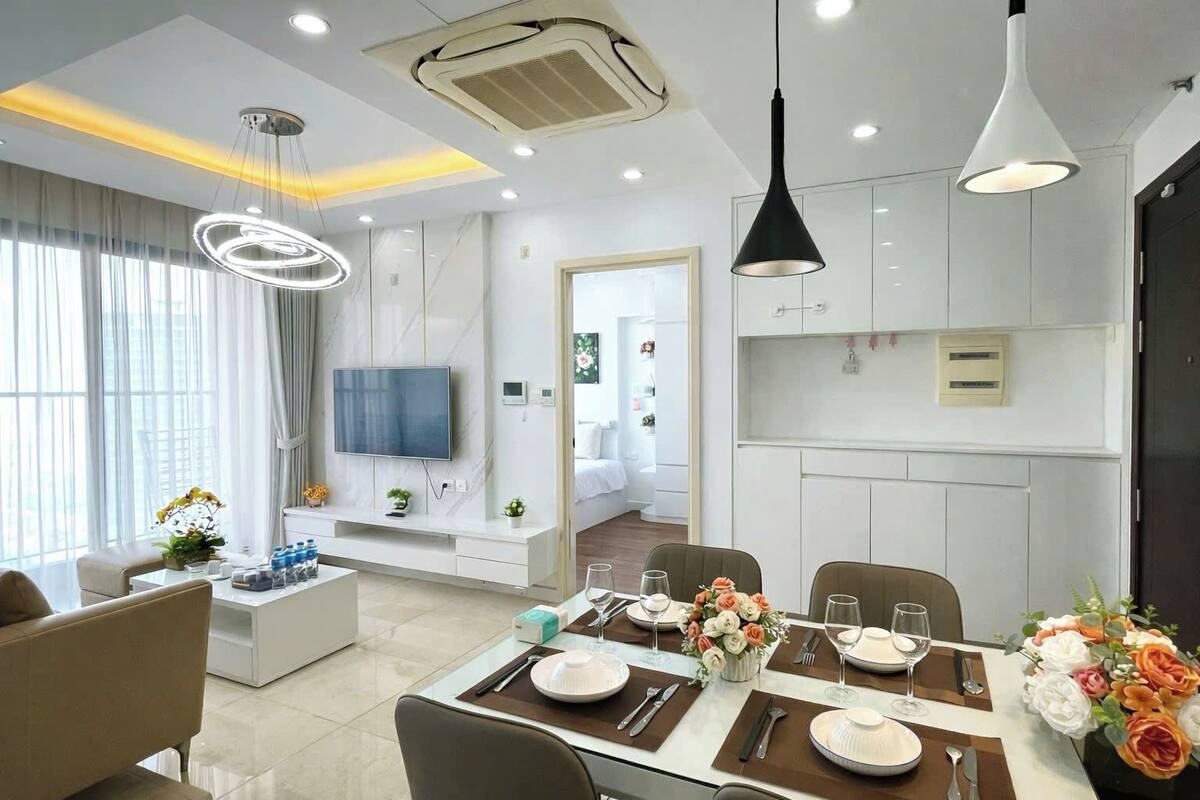 A modern living room with a dining area, a TV mounted on the wall, and a kitchen with white cabinets.